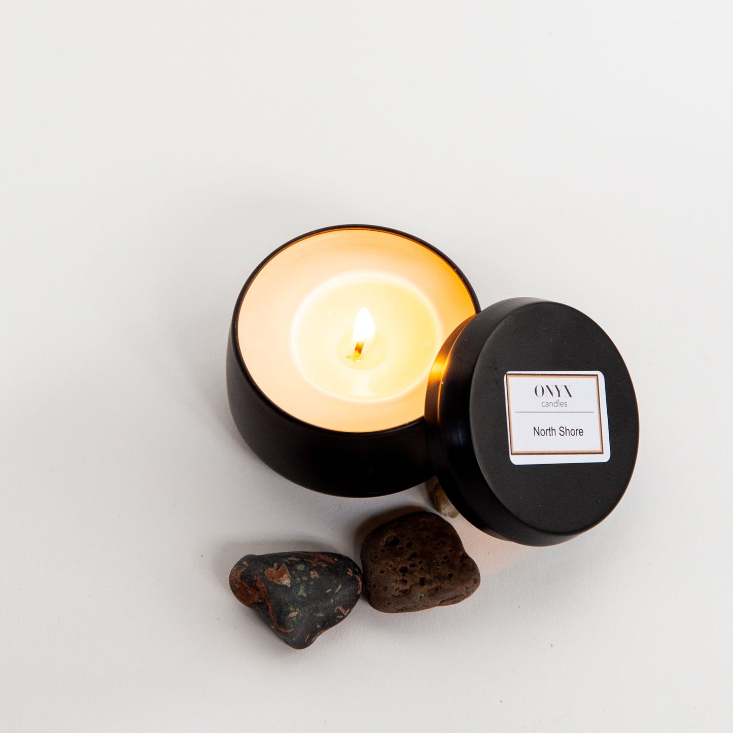 North Shore candle burning brightly in the 4oz matte black tin option. The candle is lit and surrounded by stones from Lake Superior