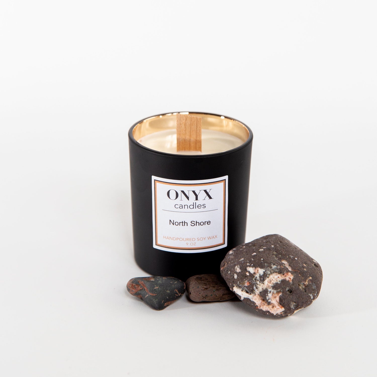 Pictured here is the 9oz matte black variant, featuring a gold interior and wooden wick. The candle is surrounded by natural stones from Minnesota's North Shore